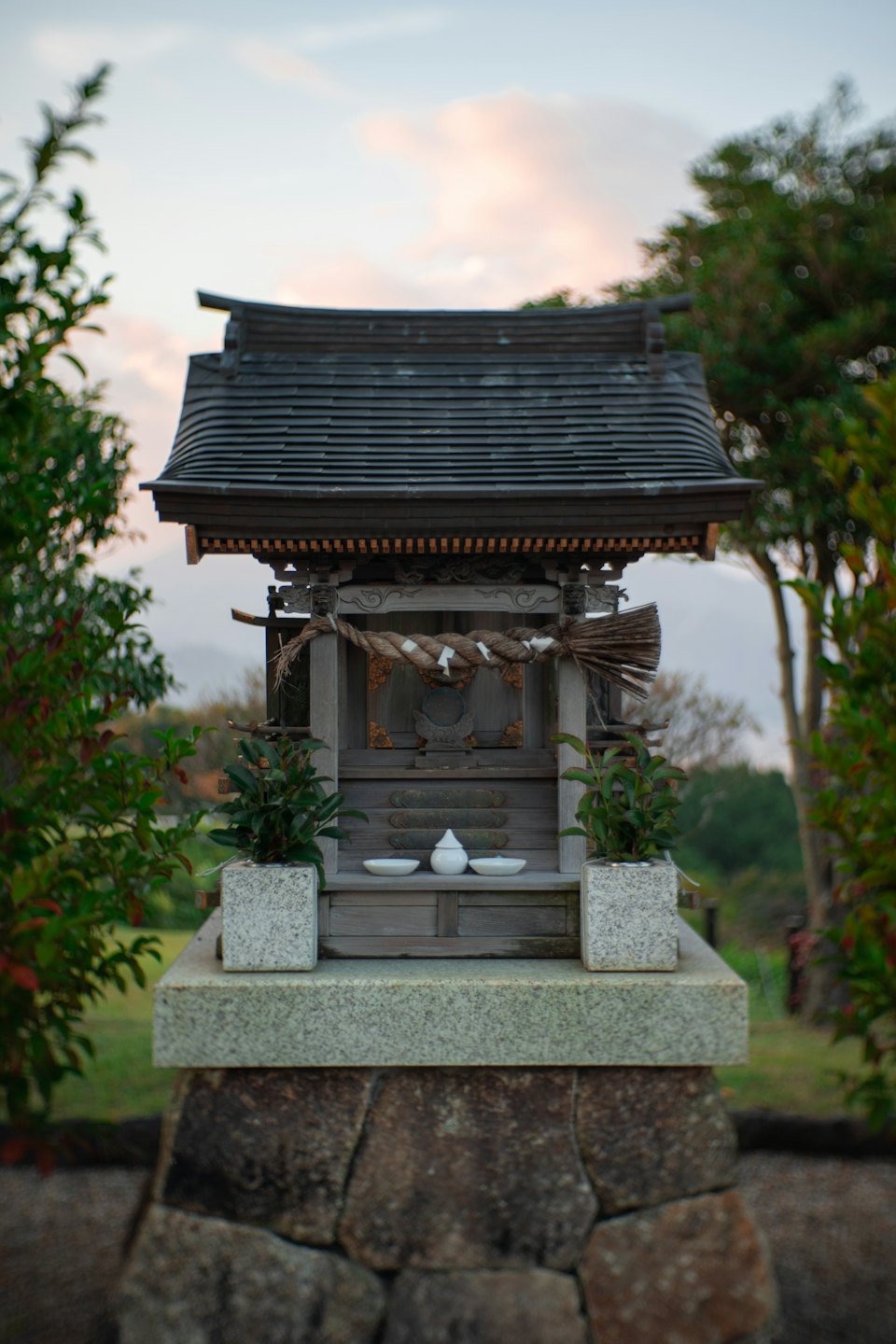 石台の上に小さな木造の日本式神社があり、お供え物が置かれています。